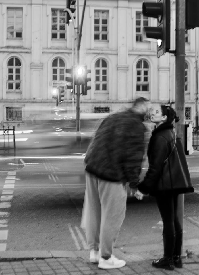 Black and white photo of a couple kissing on a city street at night, featuring motion blur and urban architecture.